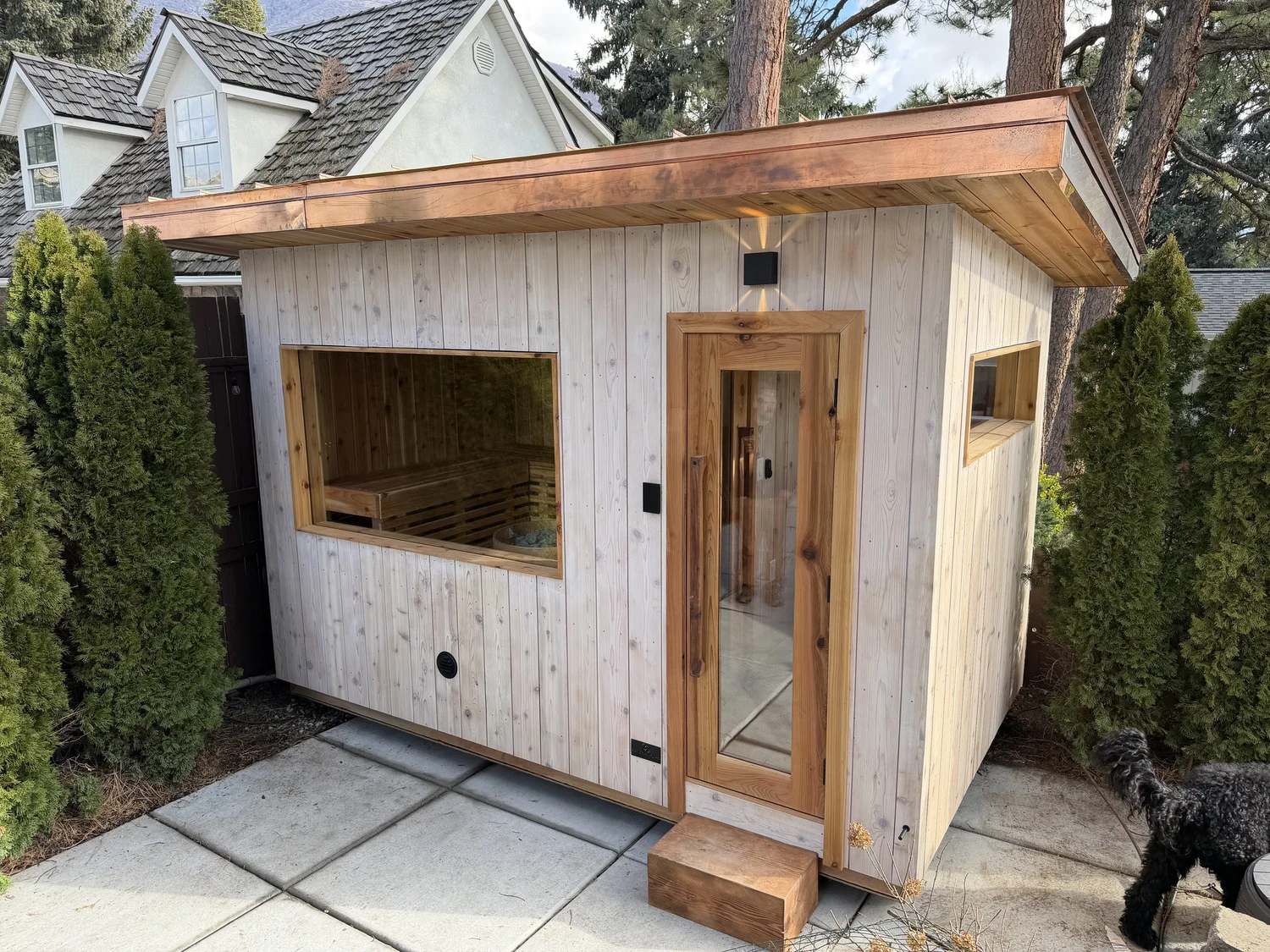 Pale whitewashed sauna with a copper roof tucked between evergreen hedges